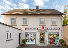 Die Außenansicht der Hinterland-Apotheke. Zu sehen ist eine Hausfront mit Schaufenstern und einem Schild mit dem Namen der Apotheke.